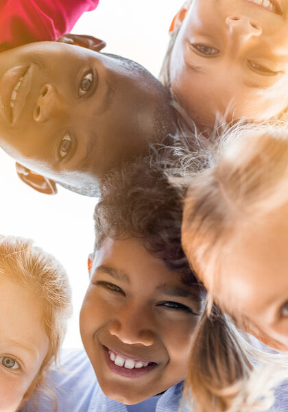 Portrait of happy kids in circle looking down and embracing. Group of five multiethnic friends outdoor looking at camera and smiling. Closeup face of children looking at camera together at park.